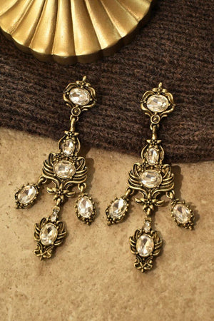Pair of antique gold chandelier earrings with clear oval crystals arranged on a textured brown fabric surface with warm studio lighting.