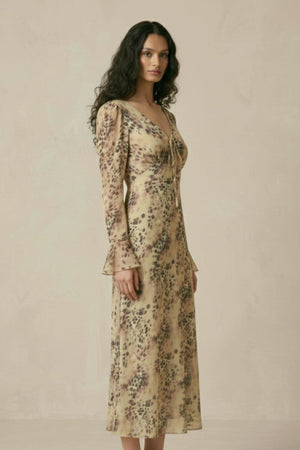 A woman standing in a beige animal-print chiffon midi dress featuring long sleeves, a tie-front neckline, and a flowing A-line silhouette, shown from the side.