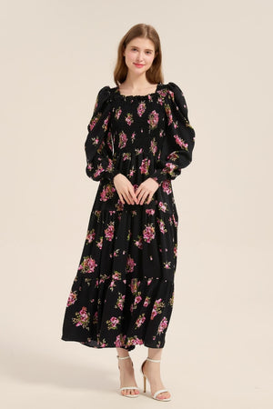 Model posing front view in a black floral smocked maxi dress with pink blossoms, puff sleeves, and a flowy tiered skirt, paired with white heels.
