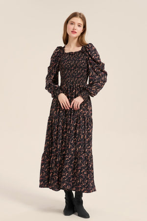 Model posing front view in a black floral smocked maxi dress with long puff sleeves and a ruffled square neckline, styled with ankle boots.