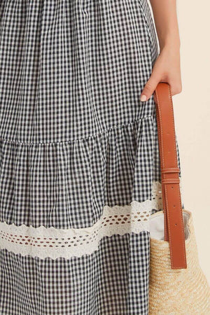 Close-up of a black gingham midi skirt with cream lace inserts and scalloped white eyelet hem, paired with a woven straw tote bag.