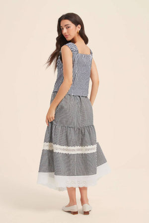 Back view of a woman wearing a black gingham top with wide straps and matching lace-trim tiered skirt, paired with white flats.