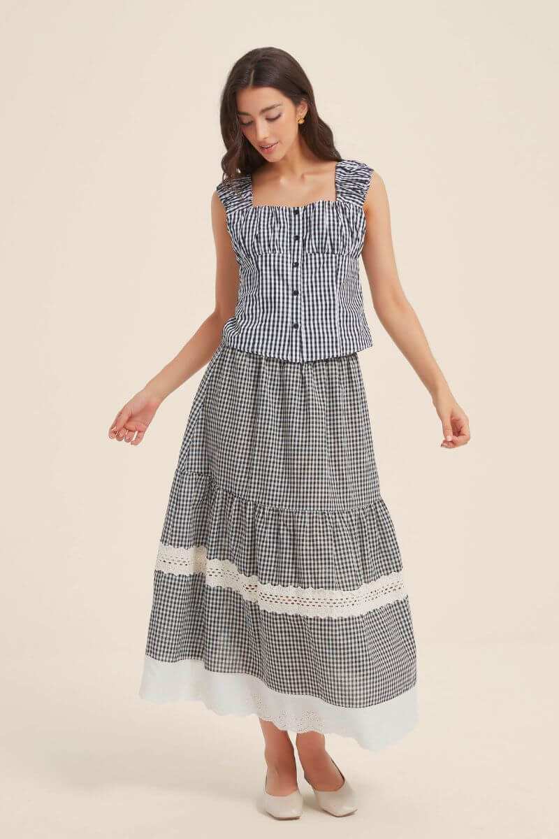 Front view of a woman wearing a black gingham two-piece outfit featuring a ruffled-strap button-front top and a tiered maxi skirt with lace trim and scalloped white hem.