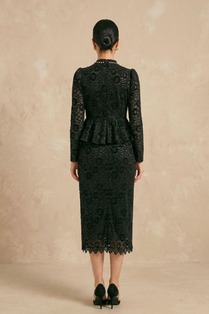 Model shown from the back wearing a black lace midi dress with long sheer sleeves, a fitted waist, peplum detail, scalloped hem, and a concealed back zipper against a soft neutral studio backdrop.