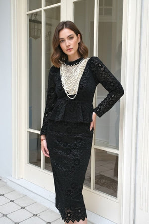 Model standing by a glass-paneled door wearing a black lace peplum midi dress with long sleeves and scalloped hem, styled with layered pearl statement necklace for an elegant vintage-inspired look.