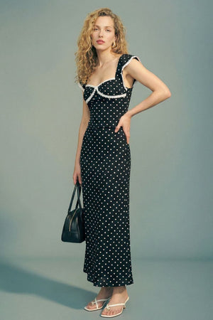 Model stands in profile wearing a black polka dot lace-trim maxi dress with a sweetheart neckline, carrying a black handbag and wearing white strappy heels in a minimalist studio.