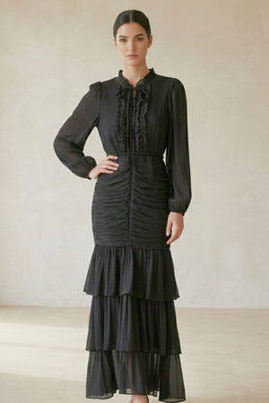 Close-up of a black long-sleeve maxi dress featuring ruffle trim, ruched bodice, and a tiered skirt, photographed against a beige background.