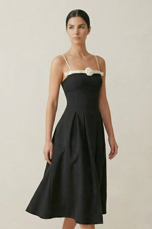 Model wearing a black midi dress with thin ivory straps and a white rosette at the neckline, styled in a clean studio setting for a refined, minimalist look.