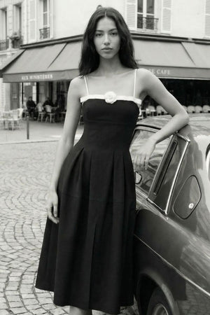 A woman leans against a vintage car on a Paris street wearing a black midi dress with thin straps, a contrast trim neckline, and a single rosette detail at the bust.