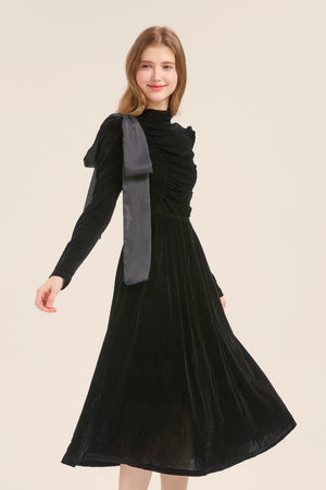Model smiling in a long-sleeve black velvet dress with ruched texture and asymmetric bow shoulder tie, styled for an evening look.