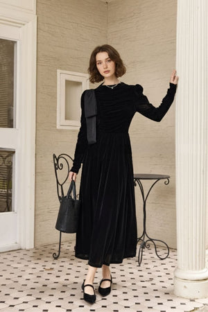 Model standing on a tiled veranda wearing a black velvet maxi dress with long sleeves, ruched bodice, and shoulder bow detail, paired with black Mary Jane heels and a bucket bag beside an iron café chair.