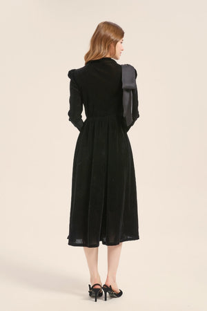 Back view of a black velvet midi dress with long sleeves and soft flare hem, featuring a large bow accent draped from the shoulder.