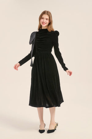 Model posing in a full-length view of a black velvet midi dress with puff shoulders, ruched front, and statement satin bow detail.