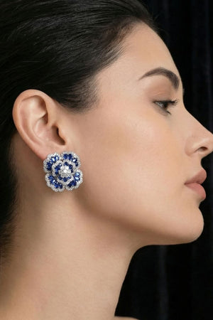 Model wearing blue floral gemstone stud earrings against a dark background, showcasing the earrings’ bold flower silhouette and jewel-toned sparkle.