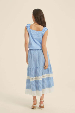 Back view of a woman in a blue gingham top with ruched shoulder straps and matching lace-insert skirt, styled with white heels and delicate gold jewelry.