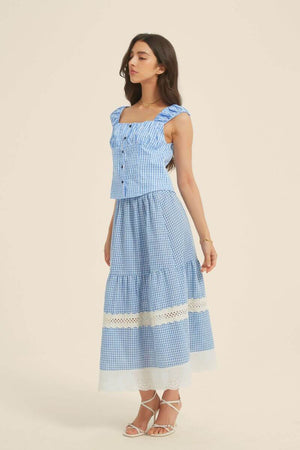 Side view of a woman in a blue gingham ruched-strap top and lace-panel midi skirt with white embroidered hem, styled with heels and gold jewelry.