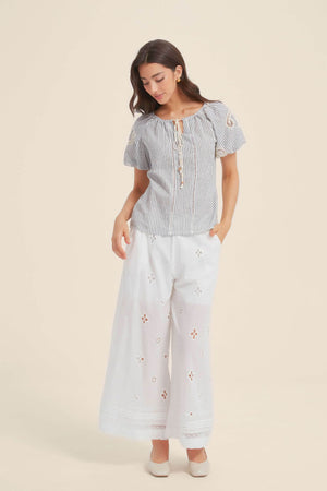 Full outfit view of a woman in a blue and white striped lace-trim blouse with tie-front tassels, paired with white eyelet wide-leg pants and cream shoes for a soft summer look.