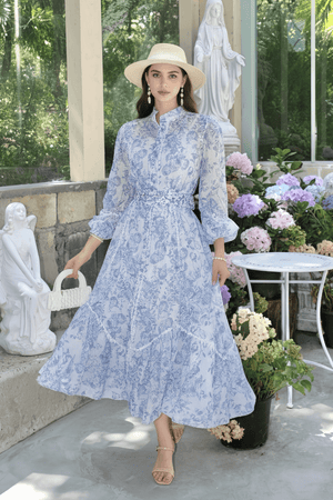 A woman in a light blue toile print puff-sleeve maxi dress with a belted waist, accessorized with a straw hat and pearl earrings, standing among hydrangeas and statues.