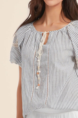 Close-up of a blue and white striped boho blouse with crochet lace inserts and tasseled tie neckline, showing lightweight cotton texture and feminine detailing.