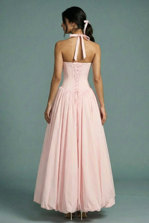 A model is shown from the back wearing a blush pink halter corset maxi dress with lace-up detailing, a soft gathered skirt, and ribbon ties at the neck, photographed against a muted studio backdrop.