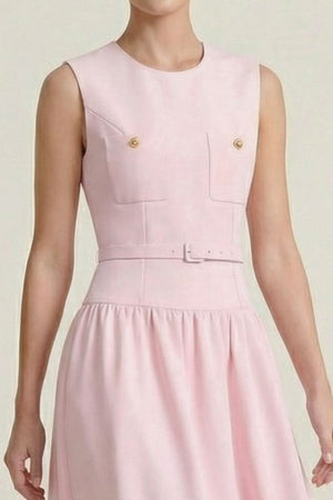 Close-up of a blush pink sleeveless dress showing the structured bodice with gold-button flap pockets and a matching slim belt in a minimal neutral studio background.