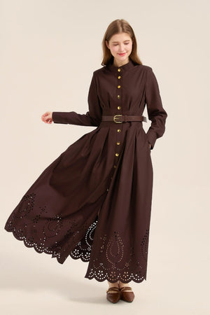 Model gracefully walking in a brown cotton long sleeve maxi dress featuring gold button-front closure, pleated waist, and eyelet embroidery hem.