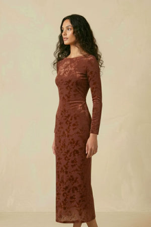 A woman standing in profile wearing a brown burnout velvet midi dress with long sleeves, a round neckline, and a slim, flattering silhouette.