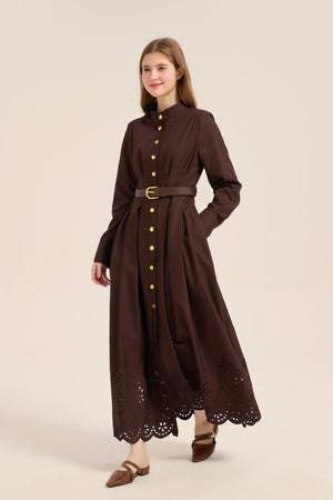 Model posing front view in a brown cotton belted shirt dress with mandarin collar, gold buttons, and laser-cut embroidered hem.