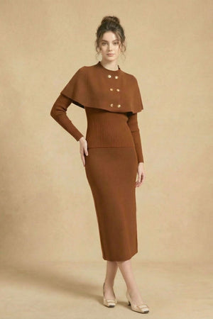 A woman stands with her hands together wearing a brown ribbed knit midi dress featuring a cape overlay and gold buttons in a neutral studio.