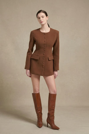 Model posing in a three-quarter angle wearing a brown tailored blazer mini dress with covered buttons and long sleeves, styled with tall brown boots in a clean white studio setting.