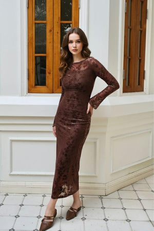 Model posing indoors against white paneled walls wearing a brown sheer velvet floral bodycon midi dress with long sleeves and boat neckline, styled with brown pointed-toe flats for an elegant autumn look.
