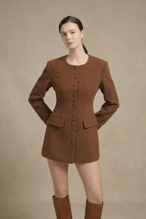 Model standing in a studio wearing a brown wool button-front blazer mini dress with long sleeves, structured tailoring, and front flap pockets, paired with knee-high brown heeled boots.