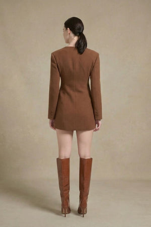 Model facing away from the camera wearing a brown wool mini blazer dress with long sleeves and structured tailoring, styled with tall brown heeled boots, photographed in a minimalist studio.