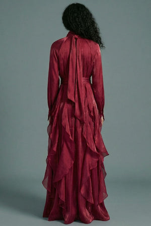 The back view of a burgundy high-neck maxi dress reveals a softly draped neckline tie and flowing ruffle layers that extend through the full-length skirt.