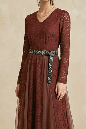 A model wears a burgundy lace V-neck midi dress with sheer long sleeves, a decorative studded belt, and a softly layered skirt against a warm neutral studio backdrop.