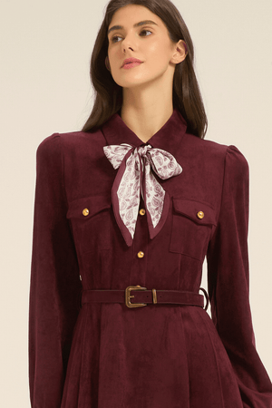 Close-up of a burgundy midi dress showing the collared neckline, gold buttons, chest pockets, belted waist, and a patterned bow scarf tied at the neck.