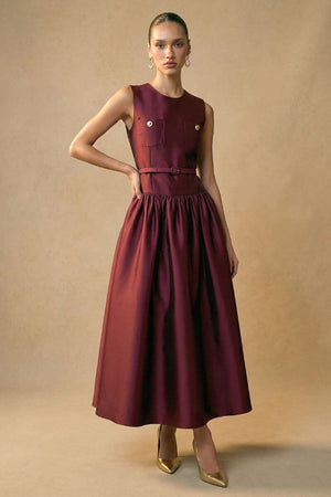 Model wears a burgundy sleeveless structured midi dress with a fitted bodice, waist belt, and softly gathered skirt, styled with gold heels against a warm studio backdrop.