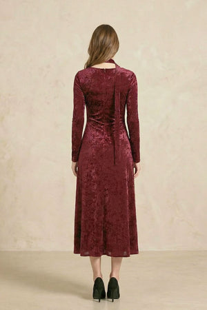 The back view of a burgundy velvet midi dress showing a clean fitted silhouette, long sleeves, a softly flared hem, and the extended velvet choker tie falling down the back.