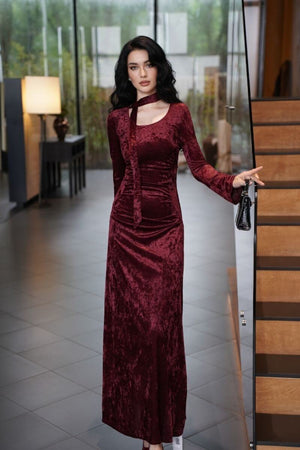 Model standing indoors near a staircase wearing a burgundy crushed velvet maxi dress with a fitted silhouette, long sleeves, and matching scarf neck tie, paired with a small black handbag for an elegant evening look.