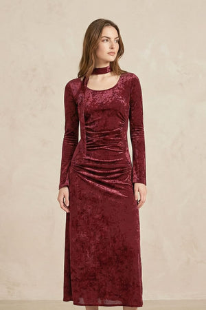 A model poses slightly angled in a burgundy velvet midi dress with long sleeves, soft draping across the waist, and a scarf-style choker neckline, creating an elegant vintage-inspired silhouette.