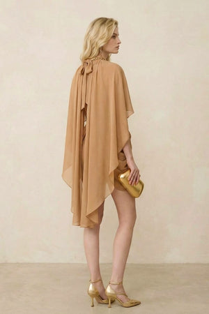 Model wearing a camel chiffon mini dress with a flowing cape overlay and gathered waist, shown from a back-side angle with soft drape movement against a neutral studio background.