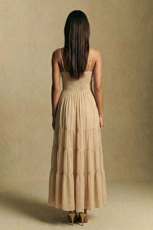 Back view of a champagne embroidered maxi dress featuring a smocked bodice, adjustable spaghetti straps, and a softly tiered skirt, photographed against a minimal beige backdrop.