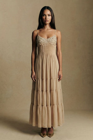 A full-length front view of a model wearing a champagne tiered maxi dress with an embroidered bust, pleated waistline, and airy chiffon layers, paired with pointed heels in a refined studio setting.