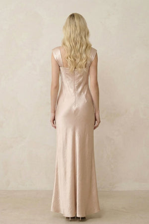 A blonde model is shown from the back wearing a champagne satin maxi dress with wide straps and a smooth, softly reflective finish against a warm neutral studio background.