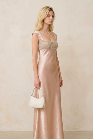 A side profile of a blonde model wearing a champagne satin maxi dress with a beaded bust, paired with an ivory shoulder bag and heels for a refined evening look.