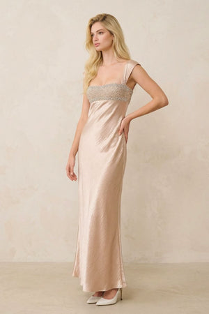 A blonde model poses in a champagne satin maxi dress featuring a beaded bust panel and wide straps, styled with ivory heels in a minimalist studio setting.