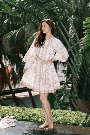 Woman wearing a white mini dress with intricate gold baroque print, belted waist, and flowy long sleeves, posing outdoors among tropical palm trees.