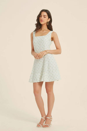 Woman wearing a light green jacquard floral mini dress with a square neckline and flared hem, styled with gold jewelry and white heels.