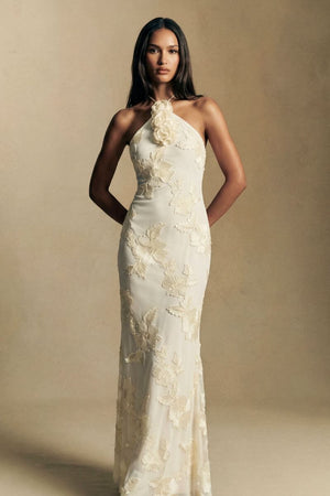 Model stands front-facing in an ivory halter maxi dress featuring tonal floral embroidery and a softly fitted silhouette, photographed in warm editorial lighting.