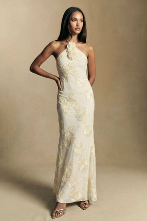 Model wearing an ivory halter-neck maxi dress with raised floral appliqué and a sculpted column silhouette, styled with delicate heels against a warm neutral studio backdrop.